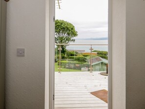 A view from a balcony with a wooden deck, a glass door, and a view of a garden and distant landscape. - 3 Moranedd, Moelfre, Anglesey. (Moelfre)
