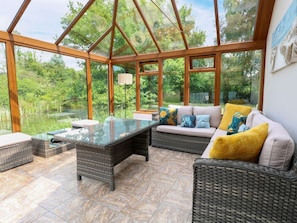 A conservatory with wicker furniture, a glass table, and a view of greenery. - Dragonfly Lodge, Whinburgh (Dereham)