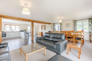 A spacious living area with a leather sofa, dining table, and chairs, a kitchen in the background, and large windows.