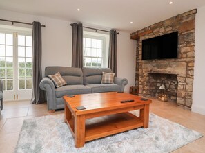 A living room with a grey sofa, a wooden coffee table, a stone fireplace, and a TV mounted on the wall. - Vine Cottage, Pilling (Preston)