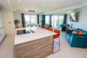 A modern kitchen with a central island, dining area with chairs, a teal sofa, and a TV mounted on the wall. - 17 Cliff Edge Newquay Cornwall (Newquay)