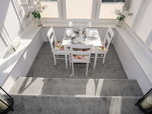 Un comedor con una mesa blanca dispuesta para cuatro personas, rodeado de sillas blancas, con vista al cielo a través de amplias ventanas.