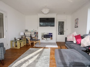 A living room with a grey sofa, a television, a bookshelf, and a window. - 5 Breeze Hill, Benllech (Tyn-y-Gongl)