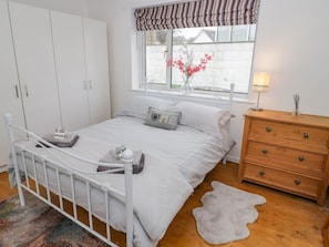 A bedroom with a white metal bed frame, a wooden dresser, a window with a view of a garden, and a bedside lamp. - 5 Breeze Hill, Benllech (Tyn-y-Gongl)