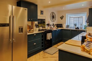 Une cuisine moderne avec un réfrigérateur en acier inoxydable, des meubles de rangement aux couleurs sombres, une horloge au mur et une fenêtre avec des stores.
