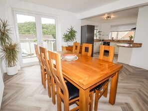 A dining area with a wooden table and chairs, a kitchen in the background, and a view of greenery outside. - 4 Bodnant Road, Rhos-On-Sea.TV (Colwyn Bay)