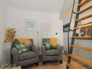 Un salon comprenant deux fauteuils, une table basse, une bibliothèque, un tableau encadré, un radiateur et un coin cuisine avec une étagère en bois.