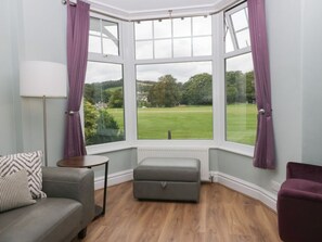 A room with a corner window, a grey sofa, a small ottoman, a lamp, and a view of a green field. - Boundary House, in Windermere. (Windermere)