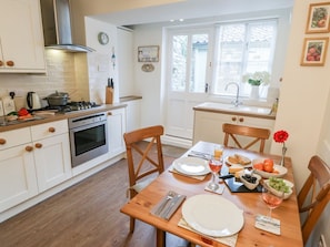 Une cuisine moderne avec des meubles blancs, une table à manger pour deux personnes et une fenêtre équipée de stores.