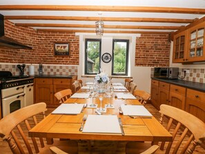 A dining room with a brick wall, wooden ceiling beams, a large window, and a dining table set for six. - Lakeside Barn, Aylsham (North Walsham)