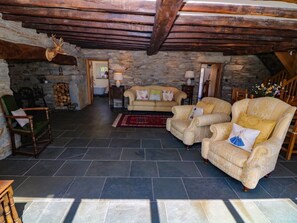 Un interior rústico con paredes de piedra, techo de madera, chimenea, sofá, sillón y lámpara.