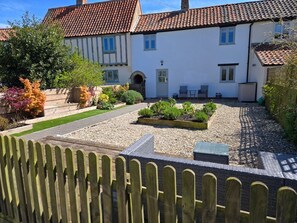 A two-story house with a tiled roof, a garden with gravel paths, and a wooden fence in the foreground. - Spring Cottage, in Sea Palling (Norwich)