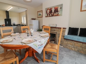 A dining area with a round table set for six, surrounded by wooden chairs. A fireplace is visible in the background, and a sofa is placed against the wall.
