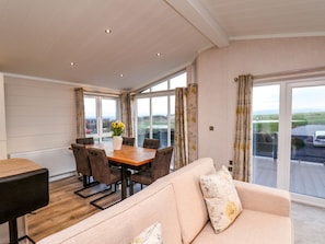 A modern living room with a sofa, dining table, and chairs, large windows with curtains, and a view of the outdoors. - Holly Lodge in North Yorkshire (Whitby)