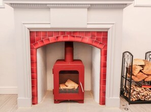 A red wood-burning stove in a fireplace with a brick arch, a stack of firewood to the right, and a metal rack with logs. - Angel Cottage, Skipton (Skipton)