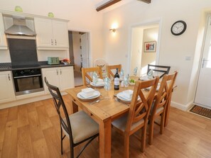 Un coin repas avec une table en bois dressée pour quatre personnes, entourée de chaises, une cuisine avec des placards blancs et une horloge au mur.