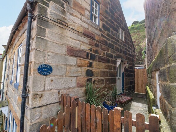 Ein Steinhäuschen mit Holzpforte, einem Schild mit der Aufschrift 'Grimes Cottage' und einem kleinen Garten mit Pflanzen.