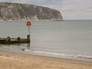Una playa con un espigón de madera, un cartel rojo y un acantilado al fondo.
