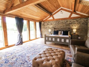 A bedroom with a stone wall, a bed, a wooden ceiling, and a large window.