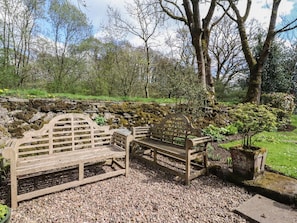 Un conjunto de banco y mesa de madera en un jardín, con un muro de piedra y árboles al fondo.