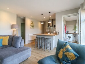 A modern living room with a grey sofa, a teal sofa with a decorative pillow, and a kitchen area with a bar stool set. - 1 Kessingland Cottages, Kessingland (Lowestoft)