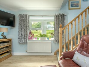 A living room with a wooden staircase, a television, a radiator, and a window with curtains. - Rose Cottage, Edlingham (Alnwick)
