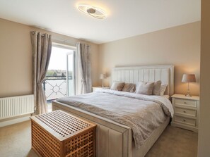 A bedroom with a large bed, a bedside table with a lamp, a window with curtains, and a wooden bench. - 21 Skippers Way, Walton-On-The-Naze (Walton on the Naze)