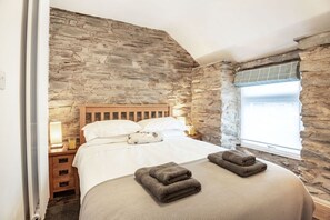 A bedroom with a stone wall, a wooden headboard, a bed with white linens, two bedside tables with lamps, and a window with a view.