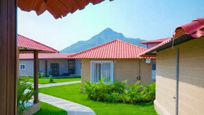 A courtyard with a pathway leading to a building with a red-tiled roof, surrounded by greenery and a mountain in the background.