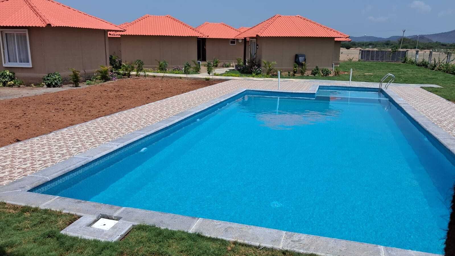 Una piscina rettangolare con bordo piastrellato, circondata da un prato e un edificio con tetti rossi.