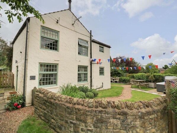 Une maison à deux étages avec un mur en pierre à l’avant, un jardin avec une table de pique-nique et un mât où est hissé un drapeau.