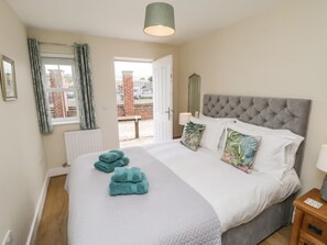 A bedroom with a large bed, a bedside table, a window with curtains, and a view of the outside. - Crossing Cottage, Whitby (Whitby)