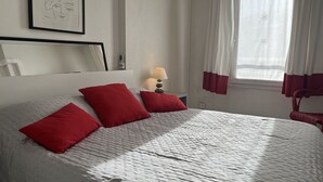 Ein ordentlich bezogenes Bett mit roten Kissen, ein Nachttisch mit einer Lampe, ein gerahmtes Kunstwerk an der Wand und ein Fenster mit Vorhängen.