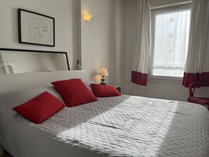 Ein ordentlich bezogenes Bett mit roten Kissen, ein Nachttisch mit einer Lampe, ein gerahmtes Kunstwerk an der Wand und ein Fenster mit Vorhängen.