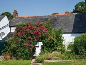 A white house with a dark roof, surrounded by lush greenery and vibrant pink flowers.