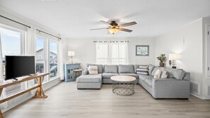Un salon moderne avec un canapé d’angle, un téléviseur posé sur un meuble en bois, un ventilateur de plafond et de grandes fenêtres avec des rideaux.