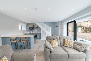 Eine moderne Küche mit einer zentralen Kochinsel, ein Essbereich mit einer Couch und Sesseln sowie eine Treppe, die in ein Obergeschoss führt.