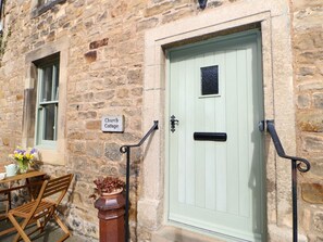 A stone building with a green door, a sign reading 'Church Cottage,' a window, and a table with chairs outside.