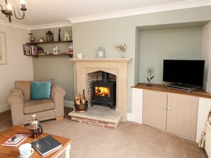 A living room with a fireplace, a sofa, a coffee table with a book and mug, a TV on a wooden stand, and a bookshelf with books and decorative items.