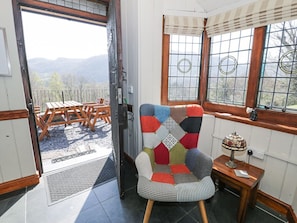 Ein Zimmer mit einem großen Fenster, einer Holztür, einem Stuhl mit Patchwork-Kissen und einem kleinen Tisch mit Lampe.