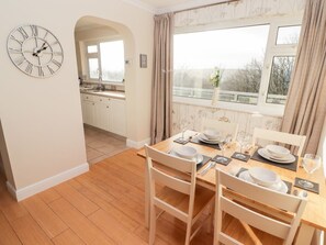 A dining area with a wooden table set for four, a clock on the wall, and a view of the outside through a window. - 54 Croft Court in Tenby, Wales (Tenby)