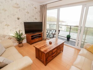 A living room with a sofa, a wooden coffee table, a TV stand, and a balcony with a table and chairs. - 54 Croft Court in Tenby, Wales (Tenby)