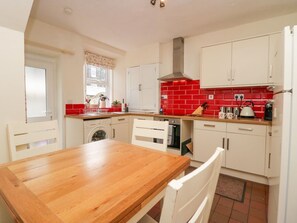 A kitchen with white cabinets, a red backsplash, a wooden table, and a window with a curtain. - Acorn Cottage is in Windermere (Windermere)
