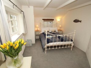 A bedroom with a white metal bed frame, a window with curtains, a bedside table with a lamp, and a vase with yellow flowers. - Sunrise Cottage, Padstow (Padstow)