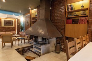 Una chimenea de ladrillo con un respiradero metálico, una estantería de madera con libros y adornos, una mesa de comedor con sillas y una zona de estar con cojines.