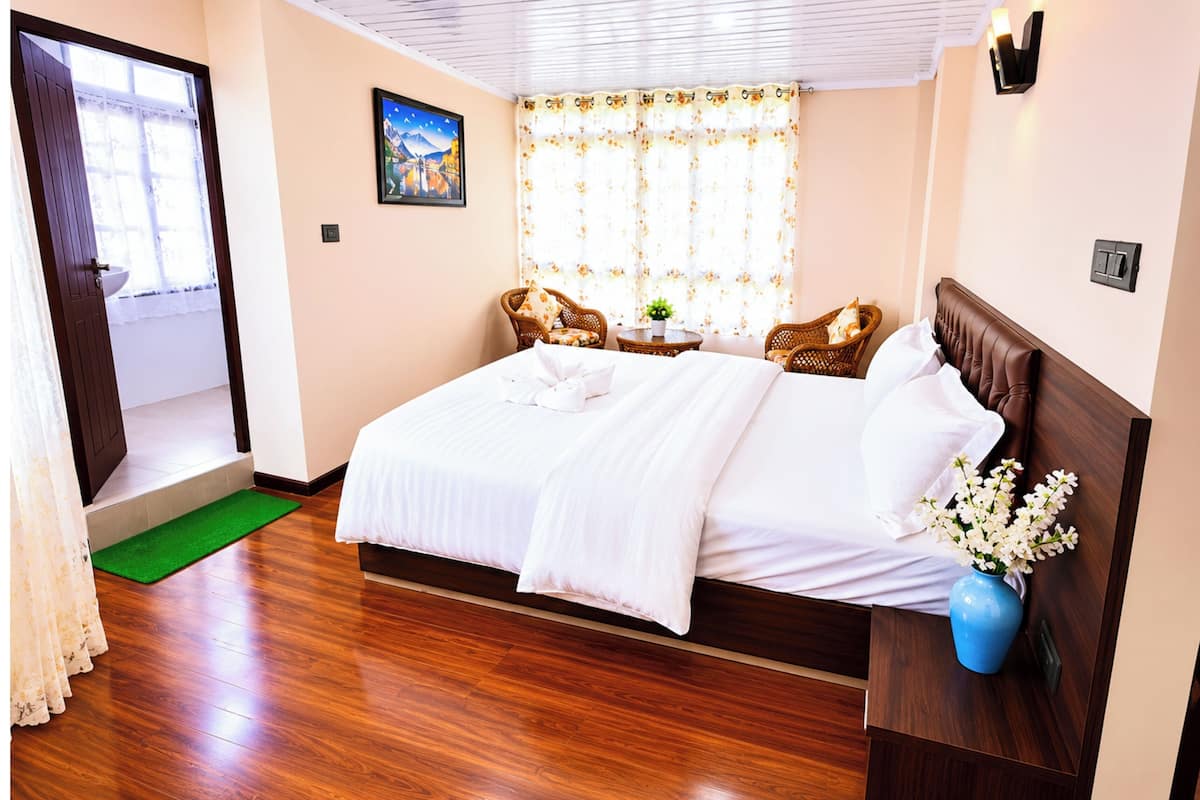 A hotel room with a wooden floor, a bed with white linens, two wicker chairs, a small table with a vase of flowers, and a painting on the wall.