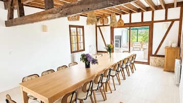 Une longue table en bois, des chaises, un plafond haut avec des poutres apparentes et de grandes fenêtres.