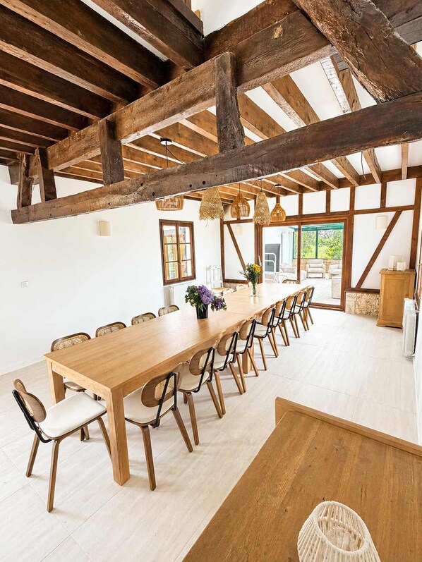 A long wooden table with chairs, a high ceiling with exposed beams, and large windows.