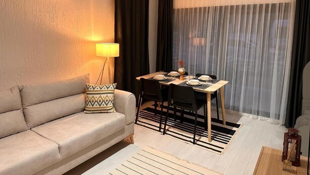 A living room with a sofa, a dining table set for four, a lamp, and a large window with sheer curtains.