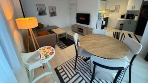 A modern living room with a TV, dining table, chairs, and a baby high chair. - ORANGE SUİTE 2+1 Family Apartment - 200m Walking Distance to the Beach (Kemer)